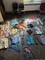 Wide layout of all girls' clothing items spread on the floor, showing variety of colors and item types including pants, shorts, and tops.