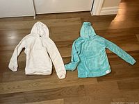Two Calvin Klein hoodies, one white and one teal, laid flat on wooden floor.