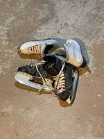 Two pairs of ice skates on concrete floor, one pair black CCM boys skates size 3 with white laces, and one pair white Jackson girls figure skates size 6 missing one shoelace.