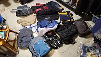 Various backpacks and bags laid out on floor including red, black, blue, and beige colors with different brands shown.