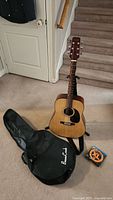 Full view of acoustic guitar placed upright on floor, black soft guitar case laying nearby, and pack of guitar strings on carpet.