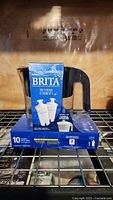 Front and side view of Brita water pitcher with box containing two Brita water filters on top, visible black pitcher handle and blue filter box with product information.