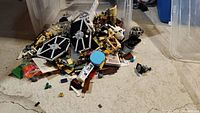 Bin of assorted LEGO pieces including Star Wars themed parts and bricks scattered on the floor with a large clear plastic bin behind them.