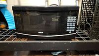 Photo of a black RCA microwave oven sitting on a wire shelf. Shown from the front to capture door and keypad detail.