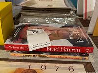 Two autographed books stacked. Top book is 'When the Balls Drop' by Brad Garrett, with a bright red spine and clear author signature. Bottom book is 'Happy Motoring! Canine Bits in the Fast Lane' showing yellow spine, signed by authors Jon Winokur and Norrie Epstein.