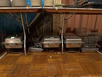 Shows five stainless steel covered chafing dishes with lids and wire racks below a folding table.