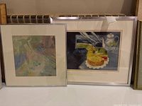 Framed monotype print showing colorful cake and teapot scene, signed by artist.