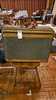 Photo of a rectangular vintage tweed fabric suitcase with leather accents and handle, placed on a wooden chair; shows nominal wear and intact zippers.