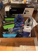 Photo showing a box containing multiple new and boxed HP and Brother printer ink cartridges and a calculator spool.