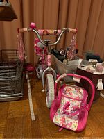Front angled view of the pink Huffy Disney Princess children's bicycle with visible training wheels, decorative handlebar streamers, and a soft pink toy baby carrier with floral patterns beside it.