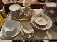 Photo showing stacked children's china plates and saucers with colorful illustrations and gold trim, small glasses with gold rims, porcelain vase, and basket dish with handle.