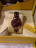 Invicta wristwatch with rose gold tone band and mauve/purple face presented in branded yellow box on cushion with tag attached.