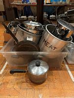 Plastic bin filled with multiple pots, pans, a cast iron pan visible at front, a large aluminum pot labeled 'MEAT', and several lids and utensils inside the bin, showing condition and contents of cooking lot.