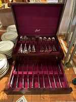 Open dark wooden silverware chest lined with burgundy velvet showing flatware in top and bottom trays, forks and spoons.