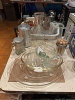 Top-down photo of vintage Glasbake clear glass meat platters, condiment bowls, pestle, glass pitchers, and small measuring cups, arranged on a table