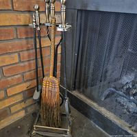 Fireplace tool set shown beside a fireplace with brick surround. Tools include poker, tongs, shovel, broom, and metal stand.