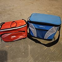 Small red Arctic Zone insulated bag and medium blue cooler bag side by side on ground.