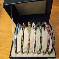 Seven multicolor cloisonné bangles displayed in a black jewelry box with white cushioned interior, showing various enamel color patterns and metal outlines.