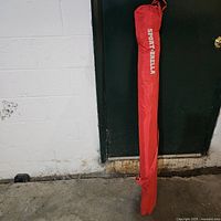 Red Sport-Brella umbrella in carrying bag standing against wall.