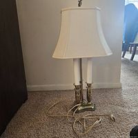 Traditional three-light table lamp with cream fabric shade, brass base, and attached cord on carpeted floor.