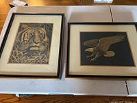 Two framed artworks side by side on a table, one depicting a tiger and one an eagle, both with black frames and dual mats.