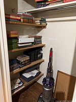 Closet shelves filled with assorted books, a Dyson cordless vacuum cleaner on the floor, and some papers and materials on the bottom shelf