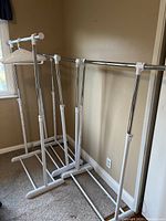 Four adjustable clothing racks standing side by side in a room next to a window. Three plastic clothes hangers are placed on one rack.