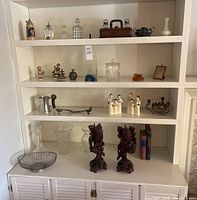 Full view of four shelves showing multiple collectibles including glassware, wood carvings, Hummel figurines, and books.