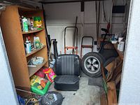 Overall view inside metal shed showing shelving unit on left, vinyl seat cushions and spare tire on floor, hand trucks and garden tools at rear