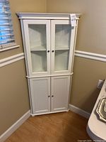 White wooden corner cabinet positioned in a corner next to window and chair, showing front closed view.