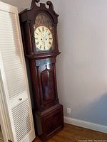 Full view of tall dark wood grandfather clock standing beside a white door. Top has curved pediment and glass front shows the clock face and paintings above the dial.