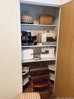 Closet view of three sewing machines on shelves with baskets above and wooden cover, stool on floor