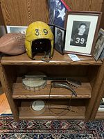 Wooden shelving unit containing all items arranged for display: yellow football helmet with number 39, vintage football, two banjos, framed photographs, and folded American flag in display case.