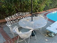 Patio set showing white metal bench, two chairs, round mesh table, and green umbrella in outdoor poolside setting.