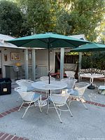 Full set of white metal patio furniture seen arranged outdoors including round table with green umbrella in center, six matching chairs with blue cushions.
