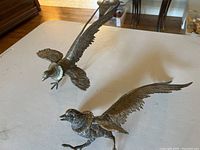 Two metal pheasant figurines displayed on a table, showing full length and wing spread details.