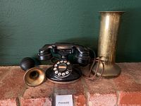 Full view of the black Western Electric rotary dial phone, two brass horn-style attachments, and tall brass cylindrical vase.