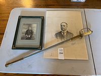 Tabletop view showing framed officer portrait, mounted gentleman portrait and sword laid across