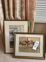 Two framed artworks leaning against curtains, one showing a house, the other showing a chair inside.