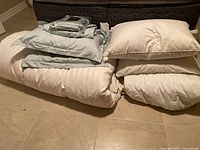 Photo showing folded bedding items including a duvet, pillows and folded sheets on tiled floor.