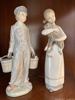 Front view of two Lladro porcelain figurines, boy with milk pails on the left and girl holding a lamb on the right