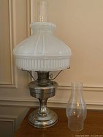Full view of the antique Aladdin oil lamp showing the frosted glass shade, tall chimney, metal pedestal base, and the spare clear hurricane chimney next to it.
