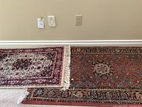 Photo showing two small area rugs placed on carpet floor, one rug on top left with rust, green, gold colors and floral patterns, the other on bottom right with red, black, cream, grey medallion pattern and fringe