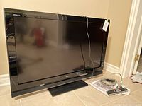 Front view of Sony Bravia KDL-40S4100 TV with stand, power and AV cables, and remote on floor next to it.