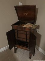 Wooden Orthophonic Victrola phonograph cabinet with top lid open revealing interior and showing some papers inside, lower cabinet doors open revealing interior framework and speaker fabric.