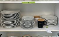 Shelf view showing multiple stacks: dinner plates on left, soup bowls center, side plates right, and two mugs with wooden lids in front
