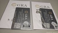 Photo showing two sets of GRA certification cards next to earrings, indicating verified moissanite oval cut stones: 1.0 carat each, total 2 carats.