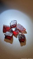 Six natural rough cherry red garnet crystals arranged on a beige surface, showing their irregular shapes and translucency.