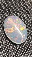 Oval cabochon Ethiopian opal gemstone on textured black background showing red, yellow and green flashes under light.