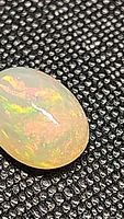 Close-up image of the oval-shaped translucent opal showing a vibrant multicolor rolling flash under clear light on textured black background.